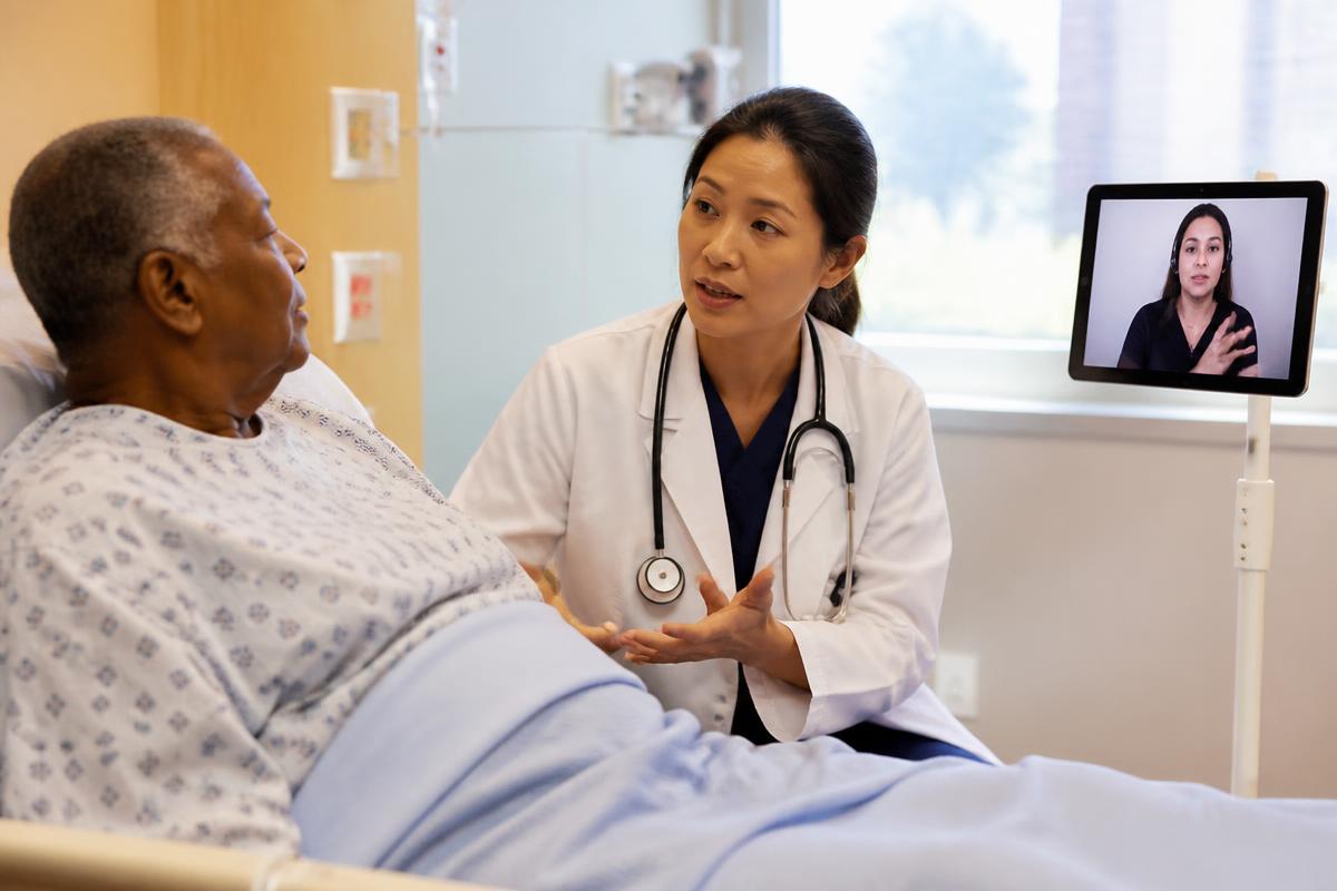 Physician speaking with a hospital patient while a remote interpreter appears on a bedside tablet